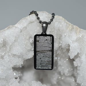 May include: A silver chain necklace with a rectangular pendant featuring a piece of meteorite encased in a black metal frame. The pendant is resting on a white crystal geode.
