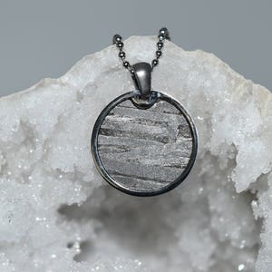 May include: A silver necklace with a round pendant featuring a slice of meteorite. The pendant is set against a white crystal background.