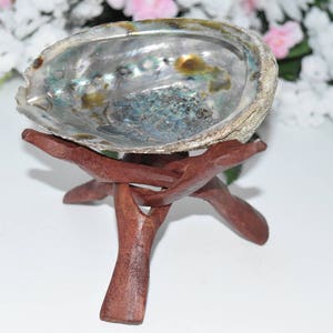 May include: An iridescent abalone shell bowl rests on a dark brown, three-legged wooden stand. The shell displays a range of colors, including blues, greens, and silver. The stand is carved with a natural, organic design.