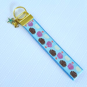 May include: A light blue key chain with a gold key ring and a starfish charm. The key chain is decorated with a pattern of brown, pink, and white seashells.