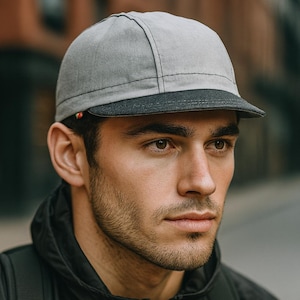May include: A five-panel baseball cap featuring a light gray crown and a dark charcoal gray brim. The cap is constructed with visible stitching along the panels and includes a small, multi-colored tab detail on the side near the brim edge.