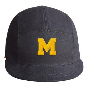 Puede incluir: Una gorra negra de cinco paneles con una letra M amarilla bordada en el centro de la parte frontal. El gorro tiene una visera plana y una corona estructurada de tela resistente, con una pequeña etiqueta naranja visible en el lateral.