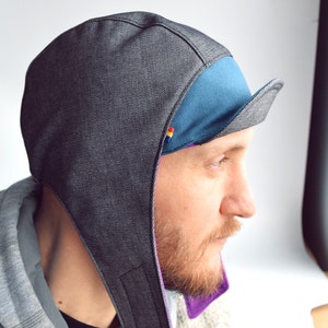 Puede incluir: Una gorra de ciclismo de denim negro con forro azul y morado y visera. La gorra tiene una solapa que se puede tirar hacia abajo para cubrir las orejas.