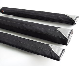 Puede incluir: Tres fundas de tela acolchada negra con un parche triangular gris plateado en cada extremo.