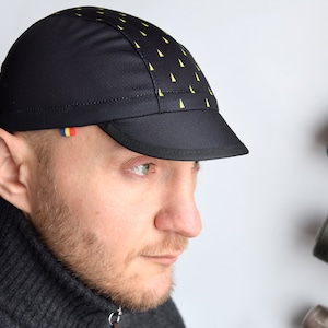 May include: A black cycling cap with a yellow and black geometric pattern. The cap has a small brim and a flag sewn on the side.