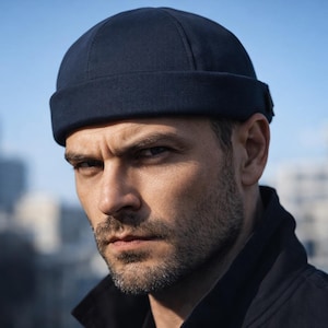 May include: A navy blue skull cap hat. The hat is made of a soft material and has a folded brim. The person in the image is wearing a black jacket. The background is blurred, with buildings and a blue sky.