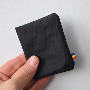 Peut inclure: Portefeuille noir avec une surface texturée et une étiquette d'accent colorée. Le portefeuille est rectangulaire et semble être fait d'un matériau durable. L'étiquette d'accentuation présente des rayures bleues, jaunes et rouges.