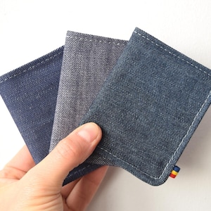 May include: Three denim cardholders in shades of blue and gray are displayed. Each cardholder is rectangular with rounded corners and white stitching. A small, colorful tag is attached to one of the cardholders.