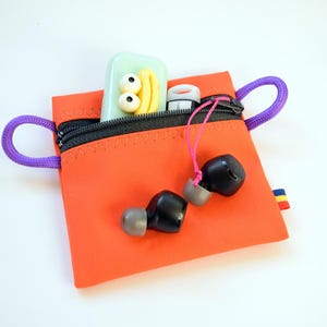 May include: An orange zippered pouch with purple rope handles, holding black earbuds, a small white and red item, and a green case with yellow and white details. Stickers of a cartoon TV and a cat are nearby.