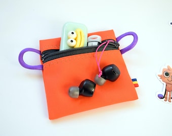 May include: An orange zipped pouch with purple rope handles, containing black earbuds, a small white and red item, and a green case with yellow and white details. Cartoon TV and cat stickers are visible.