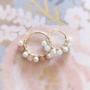 May include: A pair of gold hoop earrings adorned with small, white pearls, light blue and pink gemstones. The gemstones and pearls are attached to the hoops with gold wire. The earrings are displayed on a soft, floral background.