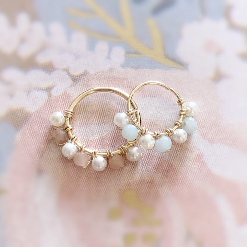 May include: A pair of gold hoop earrings adorned with small, white pearls, light blue and pink gemstones. The gemstones and pearls are attached to the hoops with gold wire. The earrings are displayed on a soft, floral background.