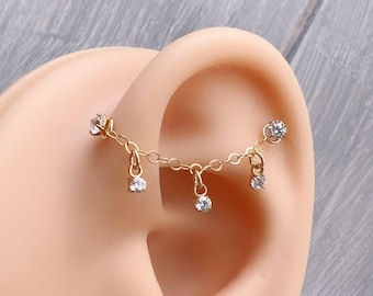 May include: Gold chain ear piercing with dangling clear cubic zirconia stones. The chain is attached to two small clear cubic zirconia stones on either side of the ear.