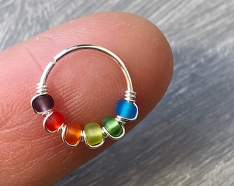 May include: A silver nose ring with a rainbow of colored beads. The beads are in shades of purple, red, orange, yellow, green, and blue. The ring is held by a finger.