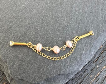 May include: A gold-colored bar pin with a chain and three pink pearls. The pin has a small clear gem on each end. The chain dangles below the pearls.