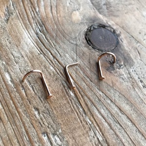 May include: Three gold nose rings on a wooden surface. The rings are all L-shaped and have a small ball at the end.