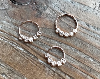 May include: Three rose gold nose rings with a cluster of small, textured beads. The rings are displayed on a weathered, wooden surface, showcasing the jewelry's design and color.