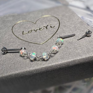 Puede incluir: Una pieza de joyería corporal plateada con una cadena y cinco cuentas de ópalo. La joyería está en una caja gris con forma de corazón y el texto "LoveYi" dentro del corazón.