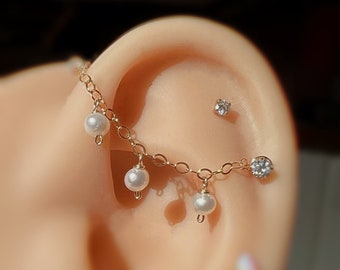 May include: A gold-colored chain earring with dangling white pearls and a small, clear gemstone stud. The earring is attached to a chain that curves along the ear, with a larger gemstone at the end.