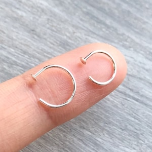 Peut inclure: Deux anneaux de nez en argent, chacun avec un petit cercle et une extrémité plate, sont montrés sur un doigt.