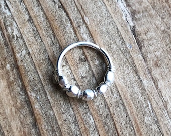 May include: A silver septum ring with a hinged design. The ring features a series of faceted beads along the front, adding texture and visual interest. The jewelry is displayed on a wooden surface.