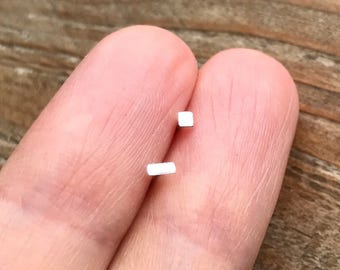 May include: Two small, silver, square-shaped earrings on a finger.