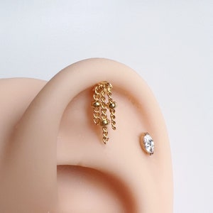 Puede incluir: Pendiente de cadena dorada con pequeñas cuentas doradas. El pendiente se lleva en un piercing de hélice.