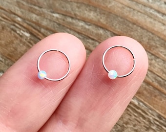 Puede incluir: Dos pequeños pendientes de aro de oro rosa, cada uno con una pequeña perla redonda de ópalo blanco. Los pendientes se muestran en primer plano, descansando sobre un dedo, con un fondo de madera.