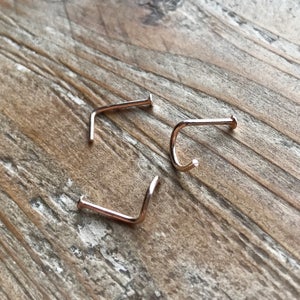 May include: Three rose gold nose rings, each with a slightly different shape. The rings are on a wooden surface.