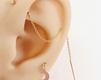 May include: Gold chain earring with a crescent moon charm hanging from the bottom. The chain is attached to a small stud earring with a clear gemstone.