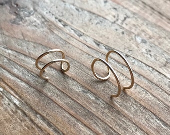 May include: Two gold-colored ear cuffs. Each cuff has a double-band design that curves around the ear. The jewelry is displayed on a wooden surface, creating a natural and minimalist aesthetic. The ear cuffs are a simple yet elegant accessory.