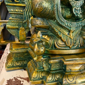 May include: A gold-colored metal statue of a deity with intricate details and a green patina. The statue is sitting on a base with ornate carvings.