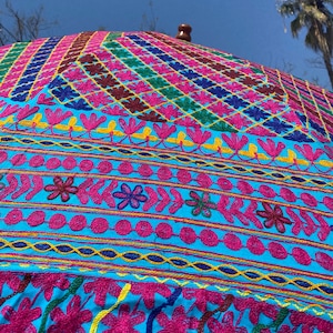 May include: A large, colorful umbrella with a blue background and intricate embroidery in pink, yellow, green, and blue. The umbrella is made of fabric and has a wooden handle.