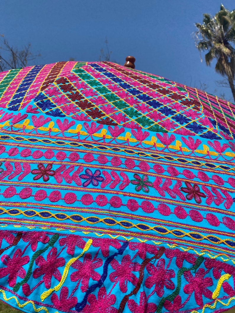 May include: A large, colorful umbrella with a blue background and intricate embroidery in pink, yellow, green, and blue. The umbrella is made of fabric and has a wooden handle.