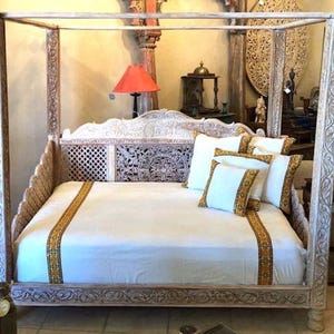May include: White wooden canopy bed with intricate carvings. The bed has a white mattress and white and gold patterned pillows. The bed is in a room with a white wall and a red lamp.