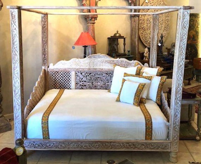 May include: White wooden canopy bed with intricate carvings. The bed has a white mattress and white and gold patterned pillows. The bed is in a room with a white wall and a red lamp.
