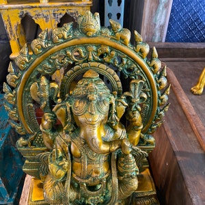 May include: A gold-toned brass statue of the Hindu deity Ganesha. The statue depicts Ganesha sitting on a throne with his trunk raised and his hands in a mudra. The statue is surrounded by a circular halo with intricate carvings.