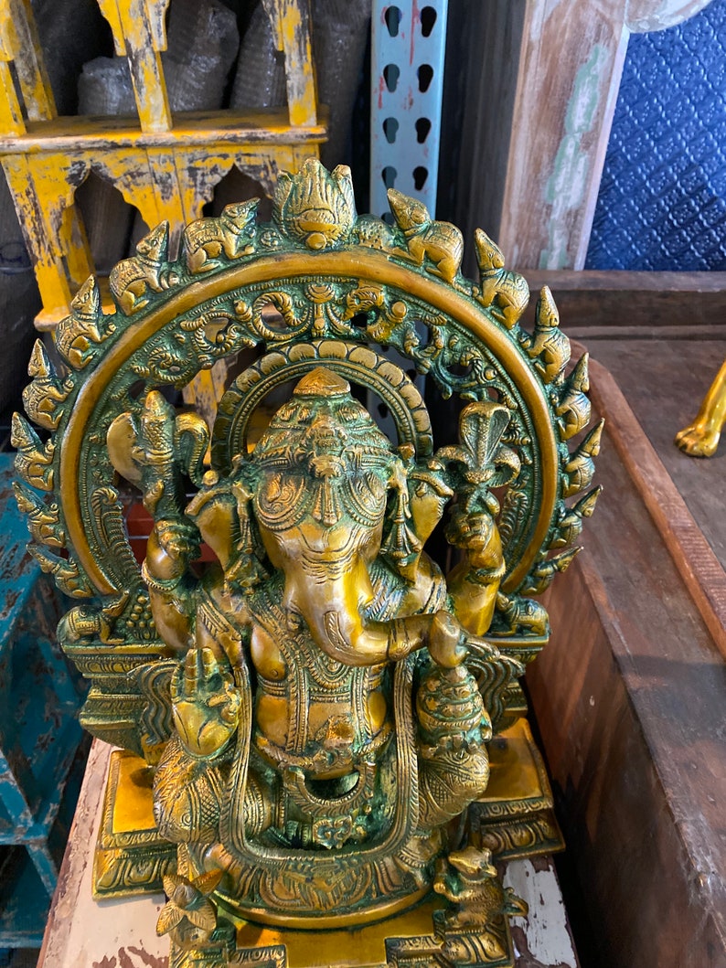 May include: A gold-toned brass statue of the Hindu deity Ganesha. The statue depicts Ganesha sitting on a throne with his trunk raised and his hands in a mudra. The statue is surrounded by a circular halo with intricate carvings.