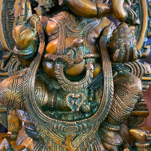 May include: A detailed, ornate, gold-toned brass statue of the Hindu deity Ganesha. The statue depicts Ganesha sitting on a throne with his trunk raised and holding a sweet in his hand. The statue is intricately carved with floral and geometric patterns.