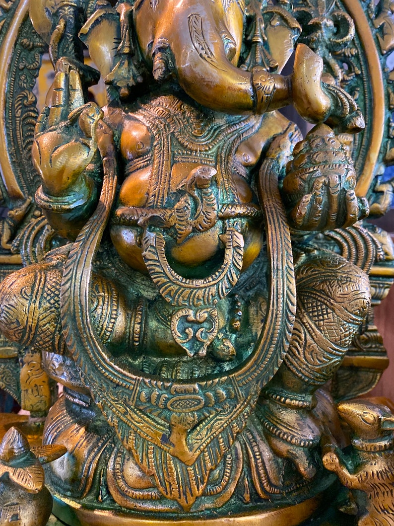 May include: A detailed, ornate, gold-toned brass statue of the Hindu deity Ganesha. The statue depicts Ganesha sitting on a throne with his trunk raised and holding a sweet in his hand. The statue is intricately carved with floral and geometric patterns.