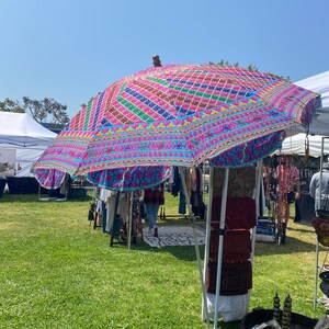 May include: A colorful, patterned canopy umbrella with a wooden handle, set up at an outdoor market. The umbrella is made of fabric with a blue background and pink, yellow, green, and red embroidery. The umbrella is providing shade for a booth selling various items, including clothing, jewelry, and home decor.