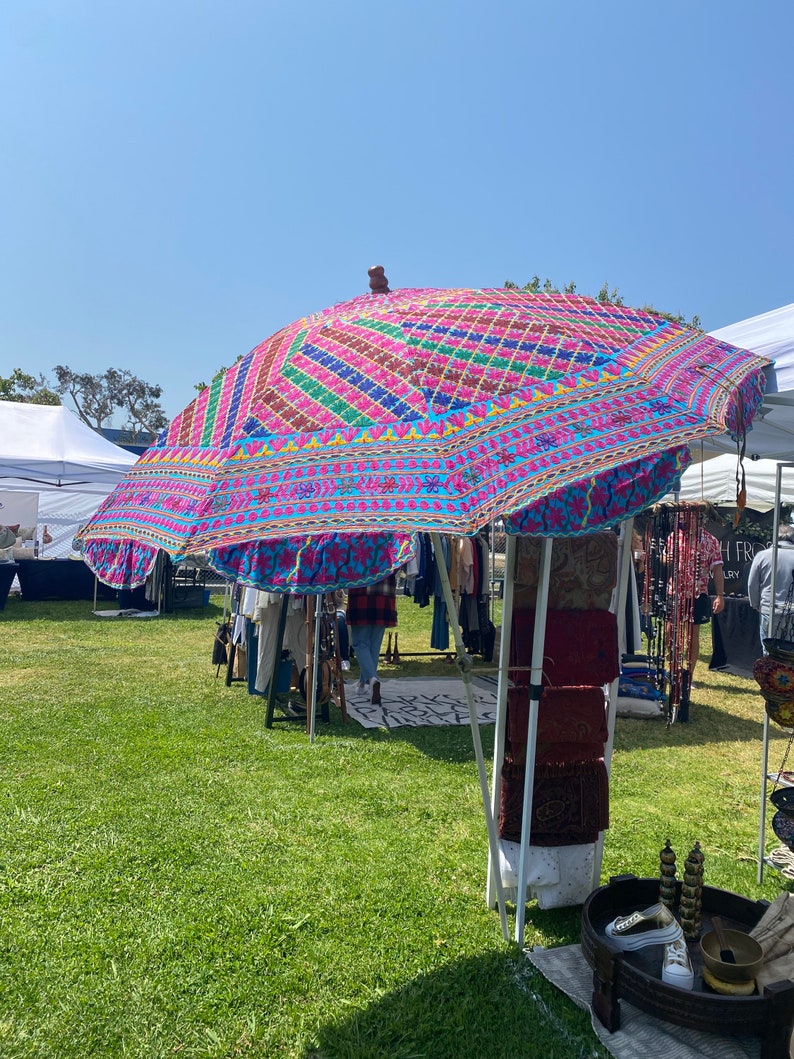May include: A colorful, patterned canopy umbrella with a wooden handle, set up at an outdoor market. The umbrella is made of fabric with a blue background and pink, yellow, green, and red embroidery. The umbrella is providing shade for a booth selling various items, including clothing, jewelry, and home decor.