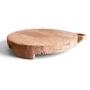 May include: A round, light brown wooden board with a weathered, textured surface. The board features a low-profile design supported by short, integrated wooden feet and includes a small, protruding handle on one side of the circular edge.