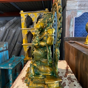 May include: A gold-toned brass statue of a deity sitting on a throne with a decorative halo behind the head. The statue has intricate details and a green patina.