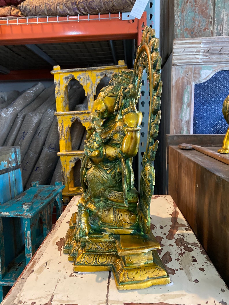 May include: A gold-toned brass statue of a deity sitting on a throne with a decorative halo behind the head. The statue has intricate details and a green patina.