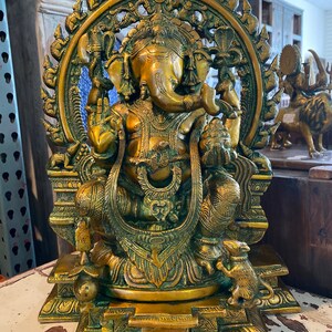 May include: A gold-toned brass statue of Ganesha, the Hindu god of wisdom and prosperity, sitting on a throne with a rat at his feet. The statue is intricately detailed and features a halo around Ganesha's head.