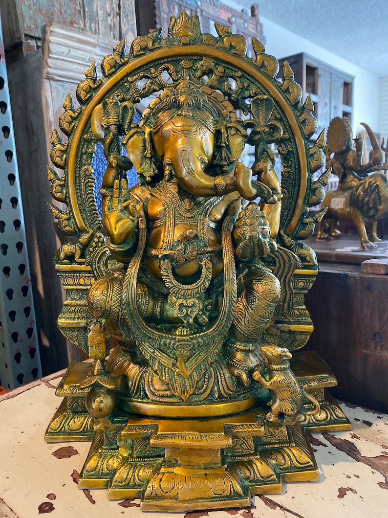 May include: A gold-toned brass statue of Ganesha, the Hindu god of wisdom and prosperity, sitting on a throne with a rat at his feet. The statue is intricately detailed and features a halo around Ganesha's head.