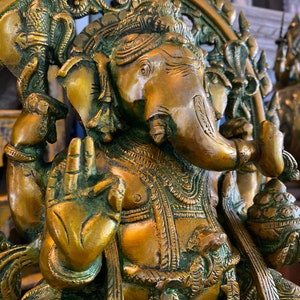 May include: A gold-toned brass statue of the Hindu deity Ganesha. The statue depicts Ganesha sitting with his trunk raised and his right hand in a gesture of blessing. The statue is intricately detailed with floral and geometric patterns.