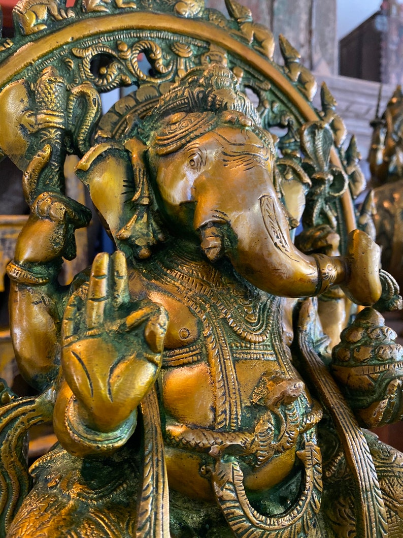 May include: A gold-toned brass statue of the Hindu deity Ganesha. The statue depicts Ganesha sitting with his trunk raised and his right hand in a gesture of blessing. The statue is intricately detailed with floral and geometric patterns.