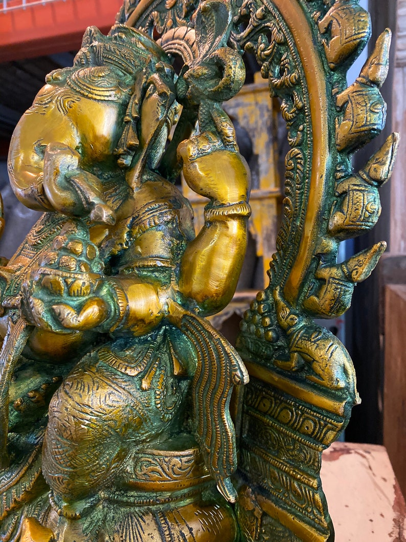 May include: A gold-toned brass statue of Ganesha, the Hindu god of wisdom and prosperity, sitting on a throne with intricate details. The statue is adorned with ornate carvings and a green patina.
